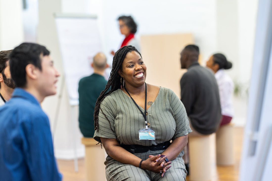 A smiling Black woman with long locs listens during a team workshop while colleagues talk and someone presents at a flip chart in the background.