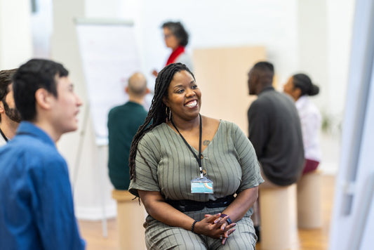 A smiling Black woman with long locs listens during a team workshop while colleagues talk and someone presents at a flip chart in the background.